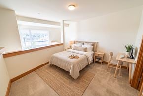 Seattle apartment for rent - Loft bedroom with carpet, a made bed, wooden nightstand, desk with monitor, and a half-wall overlooking the lower level.