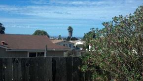 Seaside apartment for rent - A house with a brown roof is seen from behind a wooden fence with green trees in the background.