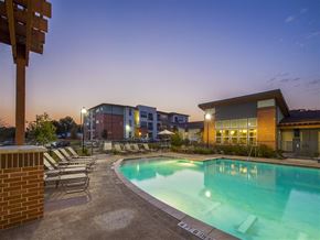 San Marcos apartment for rent - swimming pool and sundeck area at dusk featuring lounge chairs, illuminated clubhouse amenities, landscaped surroundings, and adjacent apartment buildings.