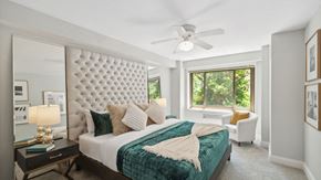 Chicago apartment for rent - Show thumbnail preview A well-lit bedroom features a large tufted headboard, a bed with teal bedding and pillows, a ceiling fan, a bedside table with a lamp, and a white armchair near a window showing