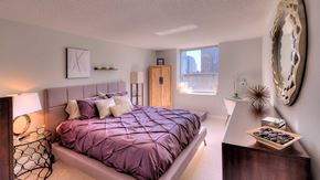 Chicago apartment for rent - Looking across a bedroom in a residence at Grand Plaza. The bed is on the left wall with lamps on both sides. The city is seen out of the window at the far end. On the right is a dresser with mirror a