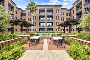 Houston apartment for rent - A patio with tables and chairs is surrounded by a brick wall and a building with windows.