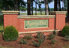 Hinesville apartment for rent - A sign for Regency Park Apartments is framed by a brick wall and bordered by shrubbery.