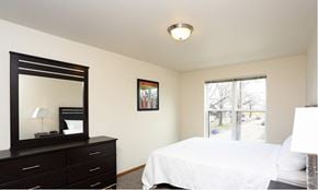Cedar Rapids apartment for rent - A bedroom with a white bed, a black dresser with a mirror, and a window with a view of trees.