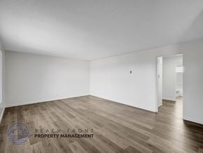 Long Beach apartment for rent - A spacious room with wooden flooring and white walls, with a logo for Beach Front Property Management in the bottom left corner.