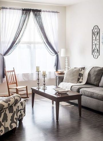 Staunton apartment for rent - A living room with a grey couch, a wooden chair, a patterned ottoman, a table with a bowl and a candle on it, and a window with curtains.