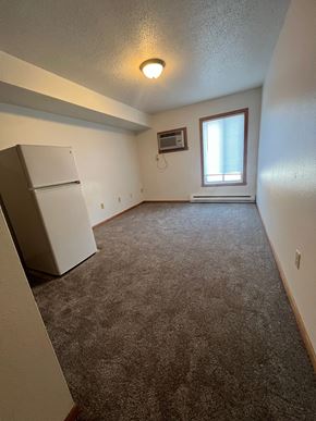 Moorhead apartment for rent - A room with a carpeted floor, a window with blinds, and a white refrigerator.