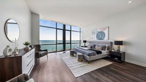 Chicago apartment for rent - Modern bedroom with large floor-to-ceiling windows offering ocean views. Features a gray bed, black nightstands, white dresser, round mirror, and contemporary art on the wall.