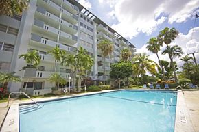 Hialeah apartment for rent - A swimming pool in front of a white apartment building with palm trees.